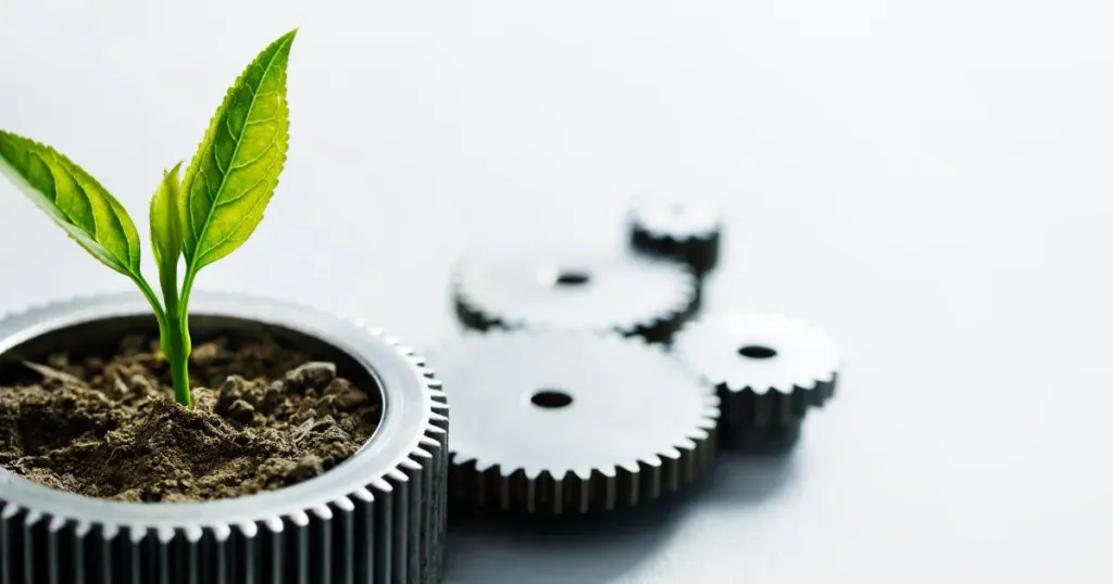 Na parte superior direita da foto, uma planta nasce dentro de uma engrenagem, que está preenchida por terra. Ao fundo, outras quatro engrenagens compõe o cenário